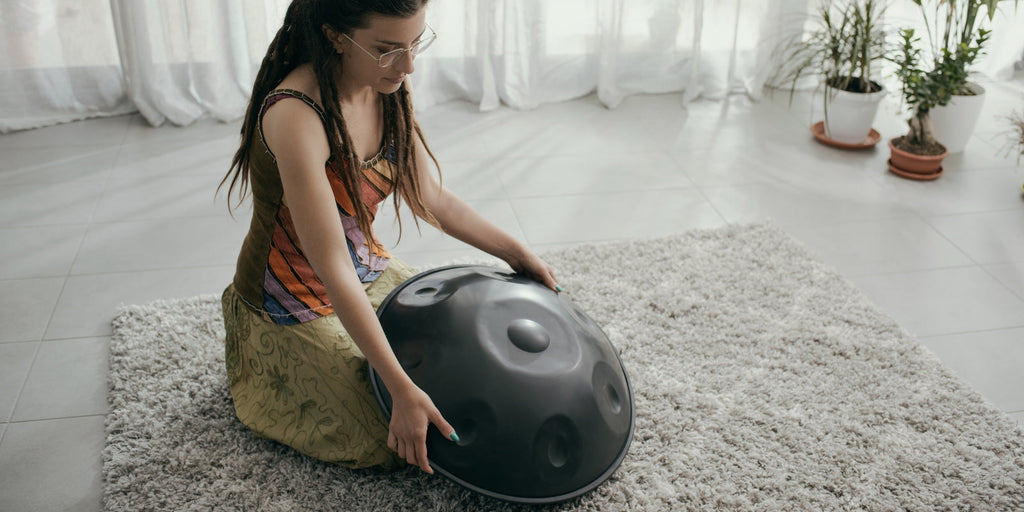 Woman kneeling on a mat holding a handpan in a calm indoor sound healing setting, illustrating a meditation instrument alternative to crystal singing bowls