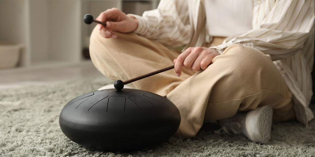 Woman sitting cross-legged playing a steel tongue drum with mallets in a calm indoor setting, illustrating a beginner-friendly meditation instrument alternative to a handpan