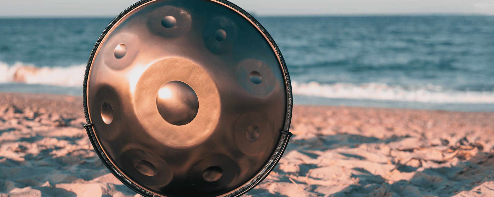 A handpan drum resting on the beach with sand and ocean waves in the background during a peaceful sound healing setting.