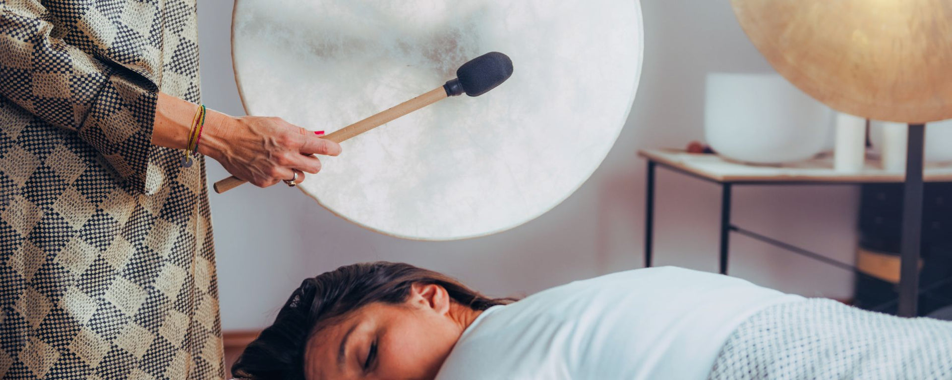 Woman playing a ritual frame drum with a mallet while another woman receives sound therapy, creating a calming and immersive healing session.