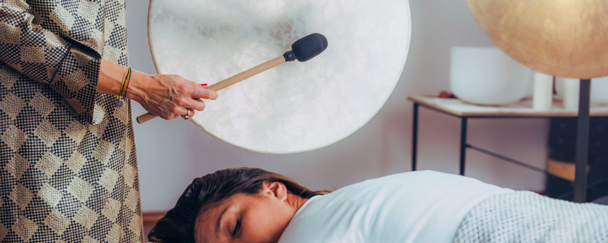 Woman playing a ritual frame drum with a mallet while another woman receives sound therapy, creating a calming and immersive healing session.