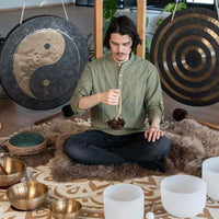 Man seated on a meditation rug holding the Meinl Sonic Energy Small Bendo Shaker SBRS featuring natural Bendo seed shells and wearable wrist-mounted design.