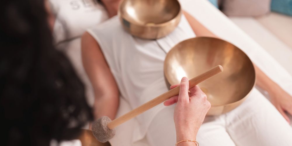Woman lying in a sound healing session with bronze alloy singing bowls placed on her body while a practitioner holds a mallet for meditation and frequency practice