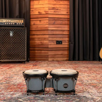 Meinl Journey Series bongo drums in Ultimate Gray (HB50UG), placed on studio floor with acoustic wood panels and amplifier in the background.