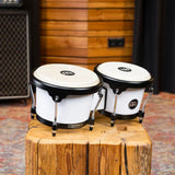 Meinl Journey Series bongo drums in white finish, positioned on a solid wood block in a studio setting, highlighting compact design, natural drumheads, and black hardware.