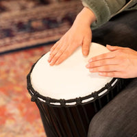 Person playing the Meinl HDJ1-M 10 inch Headliner Congo Series djembe drum, showing hand positioning on the synthetic head and rope-tuned shell in a lifestyle setting.