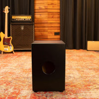 Front view of the Meinl Headliner Series Snare Cajon MCAJ100BK-CBF in a rustic studio environment, showcasing its Charcoal Black Fade finish and professional presence