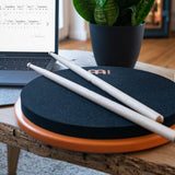 Meinl Marshmallow Practice Pad 12 inch in orange (MMP12OR), lifestyle scene with drumsticks on the pad beside a laptop and plant, illustrating quiet home practice and modern workspace use.