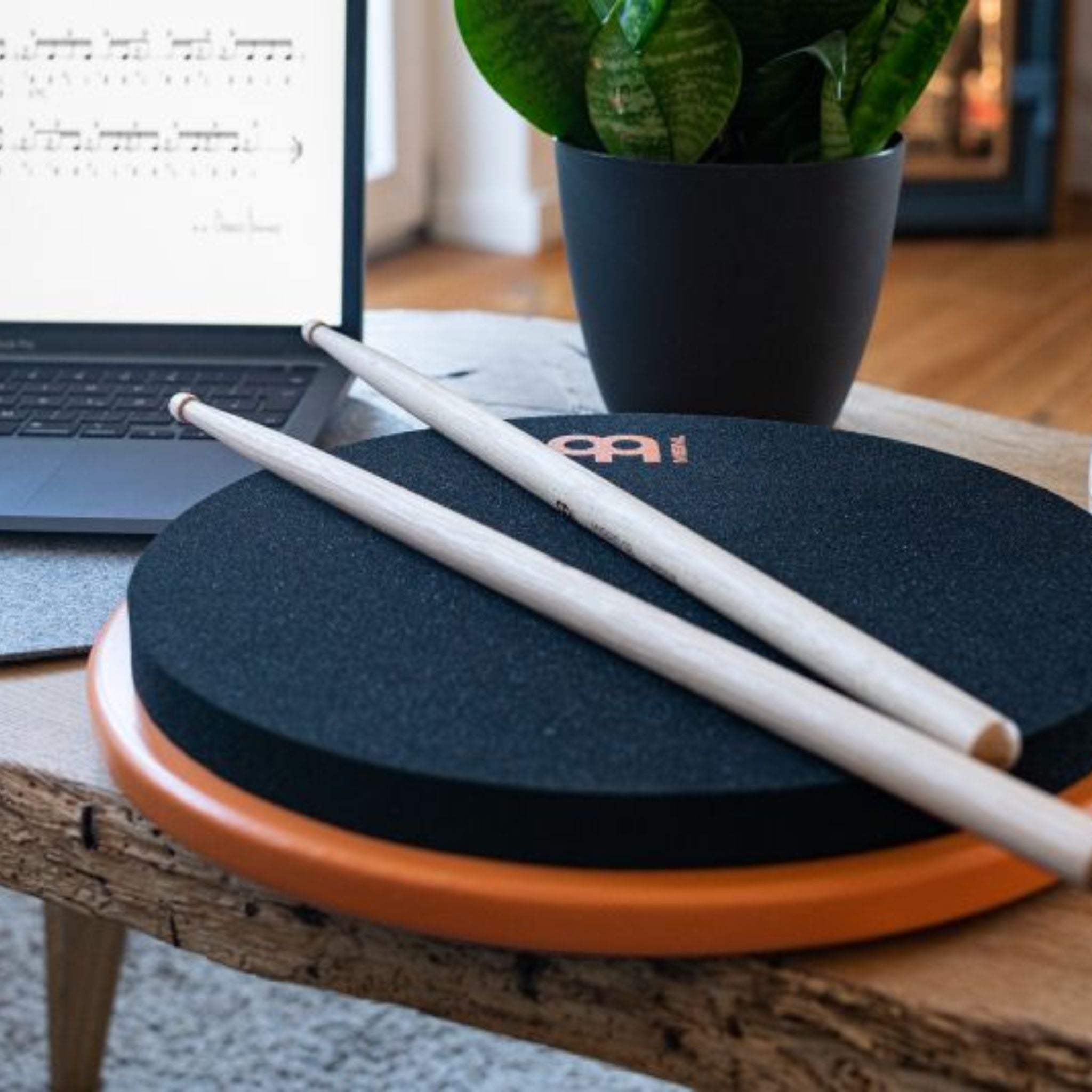 Meinl Marshmallow Practice Pad 12 inch in orange (MMP12OR), lifestyle scene with drumsticks on the pad beside a laptop and plant, illustrating quiet home practice and modern workspace use.
