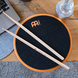 Meinl Marshmallow Practice Pad 12 inch in orange (MMP12OR), lifestyle top view with drumsticks on the pad placed on a wooden desk, illustrating quiet practice in a home or studio workspace.