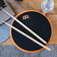 Meinl Marshmallow Practice Pad 12 inch in orange (MMP12OR), lifestyle top view with drumsticks on the pad placed on a wooden desk, illustrating quiet practice in a home or studio workspace.