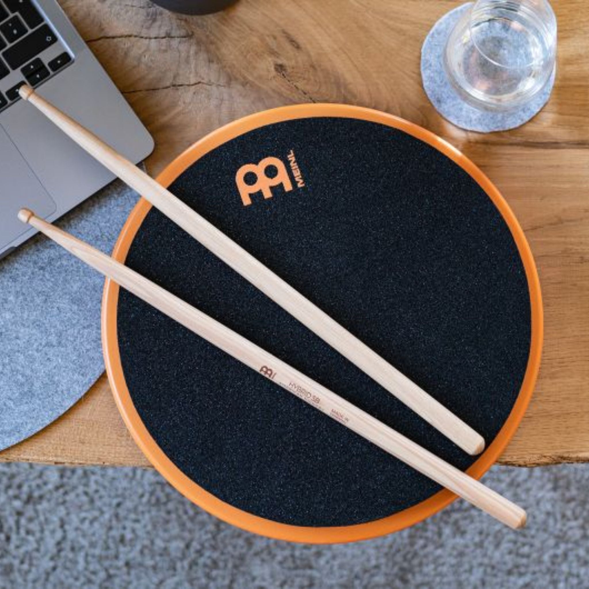 Meinl Marshmallow Practice Pad 12 inch in orange (MMP12OR), lifestyle top view with drumsticks on the pad placed on a wooden desk, illustrating quiet practice in a home or studio workspace.