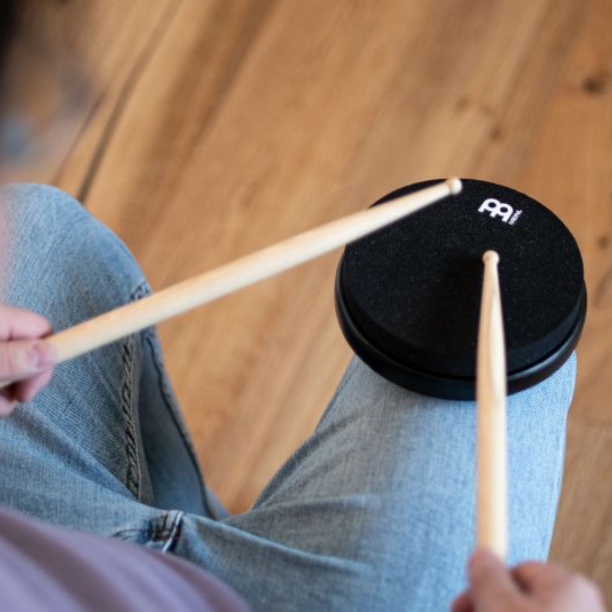 Person playing the Meinl Marshmallow Practice Pad 6" Black MMP6BK on their knee with a pair of drumsticks.