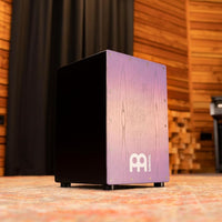 Meinl Snare Cajon Headliner Series Lilac Purple Fade MCAJ100BK-LPF in a rustic studio setting — premium percussion drum for professional performance, recording, and creative rhythm.
