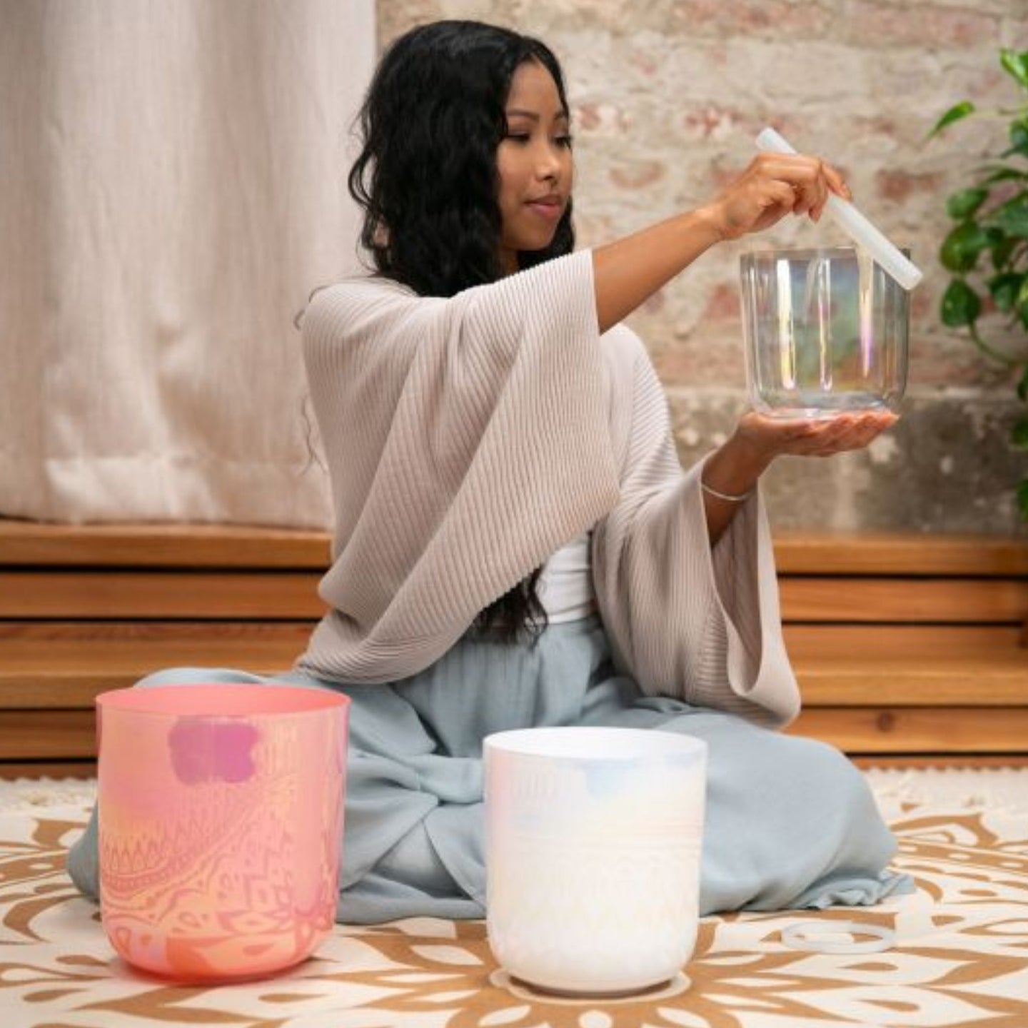 woman holding and playing the 6 inch E4 Clear Solar Plexus Crystal Singing Bowl from the Meinl Sonic Energy 3-Piece Essence Crystal Singing Bowl Set ECSBSETEBG with the B3 and G3 bowls arranged on a meditation rug for sound therapy and mindfulness practice.
