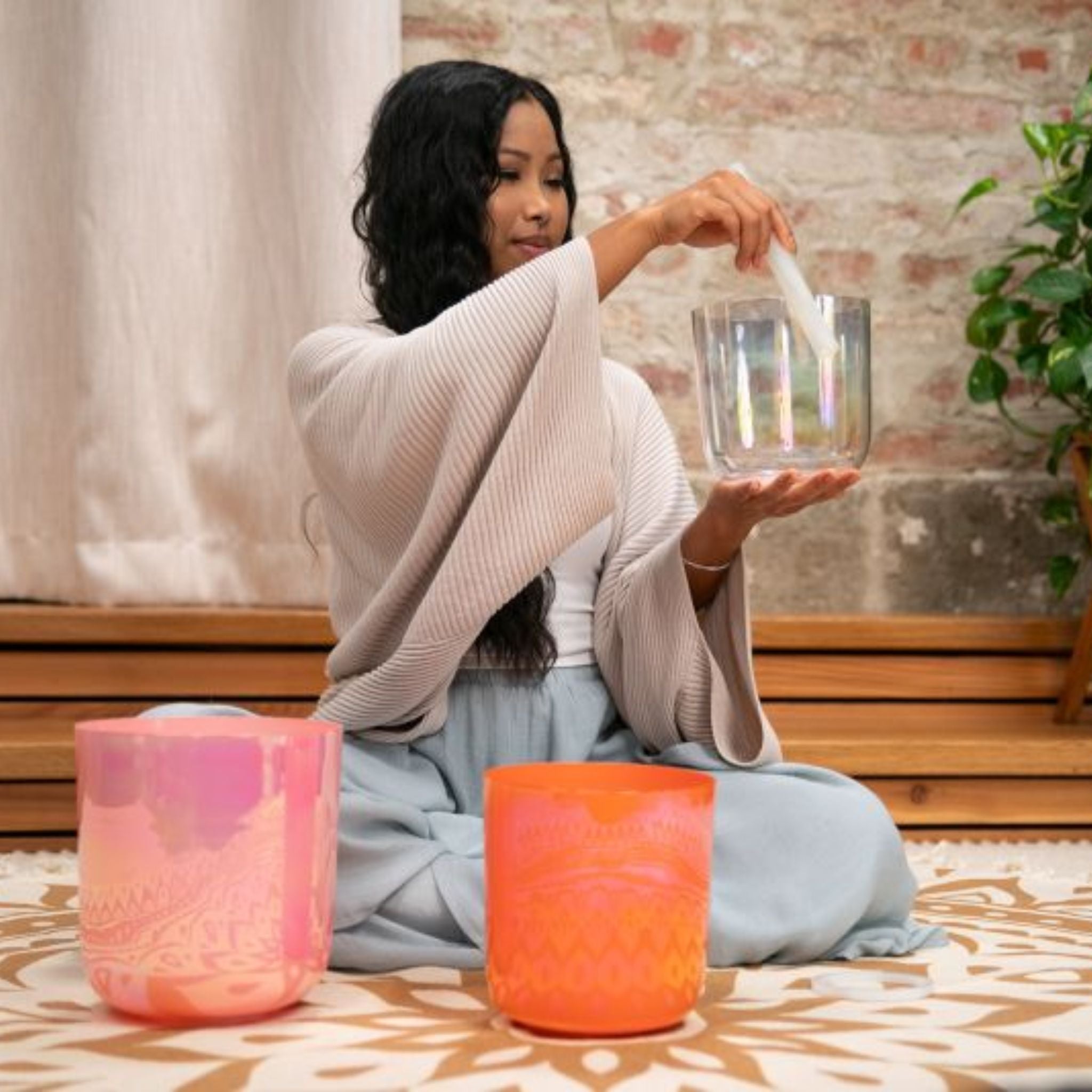 woman holding the 6 inch E4 Clear Solar Plexus Crystal Singing Bowl from the Meinl Sonic Energy 3-Piece Essence Crystal Singing Bowl Set ECSBSETECG with the C4 and G3 bowls arranged on a meditation rug in front of her for sound therapy and meditation practice