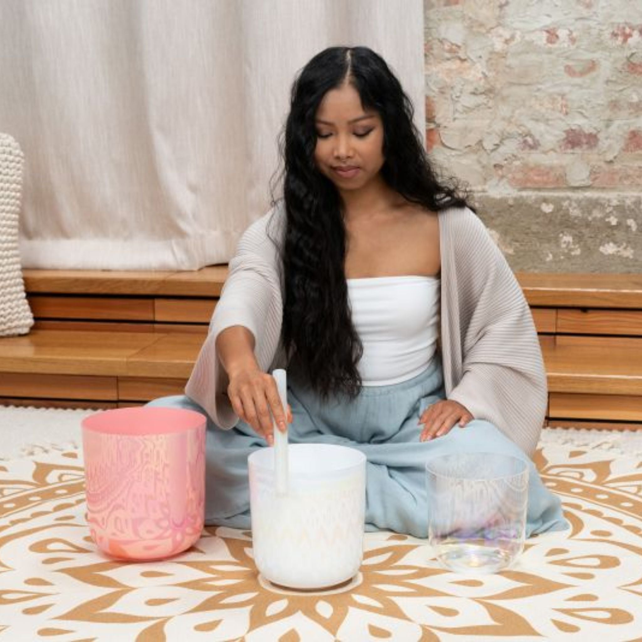woman playing the Meinl Sonic Energy 3-Piece Essence Crystal Singing Bowl Set E4 B3 G3 440Hz ECSBSETEBG on a meditation rug, creating harmonic quartz crystal tones and long-lasting resonance for meditation, sound therapy, and immersive sound exploration.
