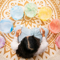 aerial view of woman playing Meinl Sonic Energy 7-Piece Marble Crystal Singing Bowl Chakra Set 432Hz MCSBSETCHA arranged on floor mat showing the full set of chakra-coloured quartz crystal singing bowls for meditation and sound therapy