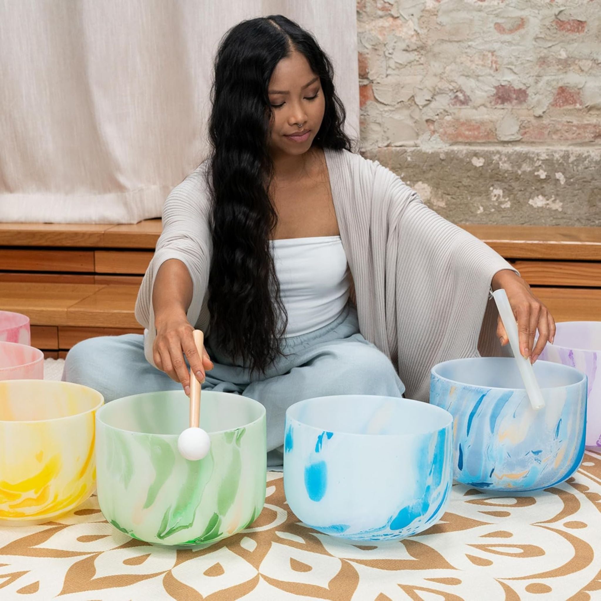 front view of woman playing Meinl Sonic Energy 7-Piece Marble Crystal Singing Bowl Chakra Set 432Hz MCSBSETCHA showing chakra-coloured quartz crystal singing bowls arranged for meditation and sound therapy