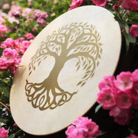 Meinl Sonic Energy Frame Drum 16 inch Tree of Life HD16WB-TOL displayed outdoors among pink flowers showing natural goat skin head and engraved Tree of Life design