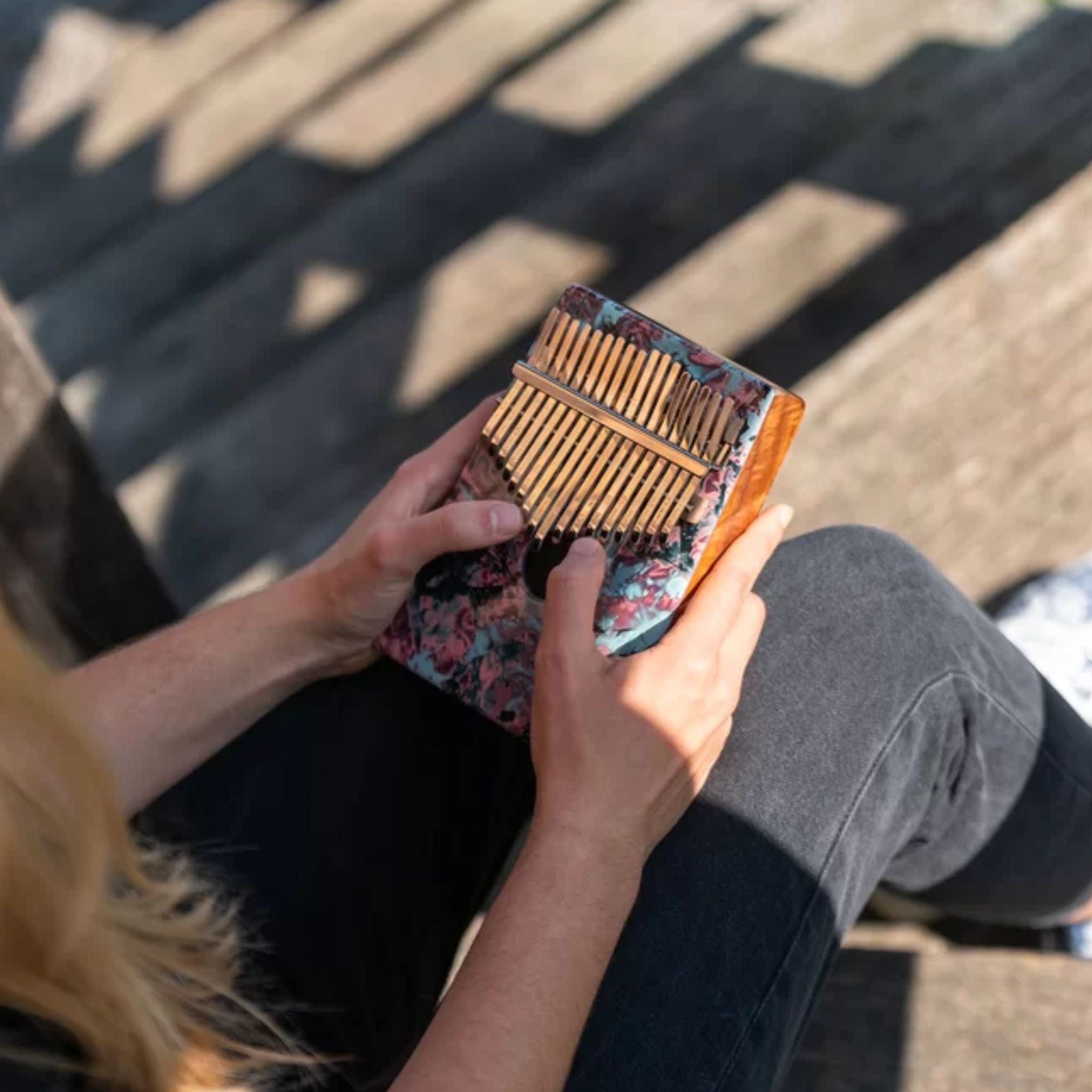 over-the-shoulder view of woman playing Meinl Sound Hole Designer Kalimba Coral Reef KA17CR showing the face of the kalimba, engraved steel tines, and coral reef artwork while creating melodic and resonant tones.