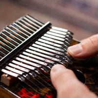 extreme close up of two thumbs playing the Meinl Sound Hole Designer Kalimba Lava KA17LV showing engraved steel tines and precise finger technique used to create melodic and resonant kalimba tones.