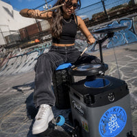 Woman playing the NUVO Zip-Kit Essential compact acoustic drum kit at a skate park with graffiti walls in the background.
