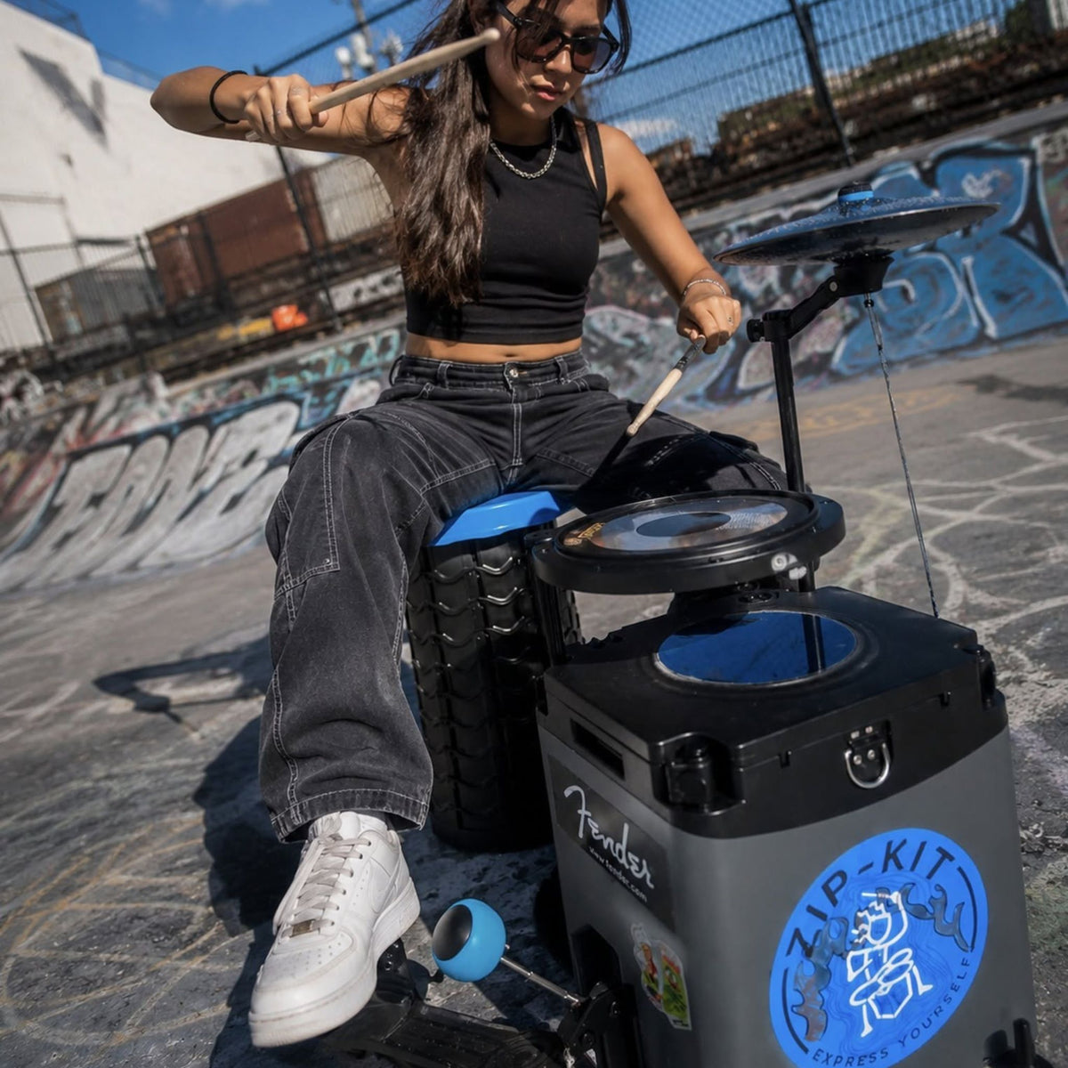 Woman playing the NUVO Zip-Kit Essential compact acoustic drum kit at a skate park with graffiti walls in the background.